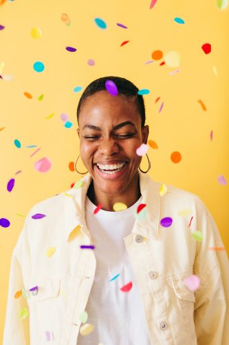 Portrait of a young woman laughing with colored confetti in front of a yellow wall.