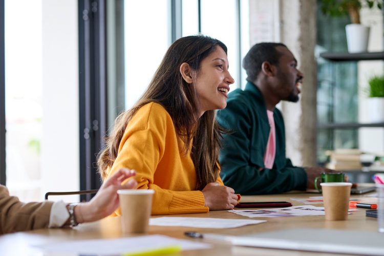 Businesswoman in casual clothes with long hair smiling and listening to colleague while sitting at table against window during business seminar in light office.